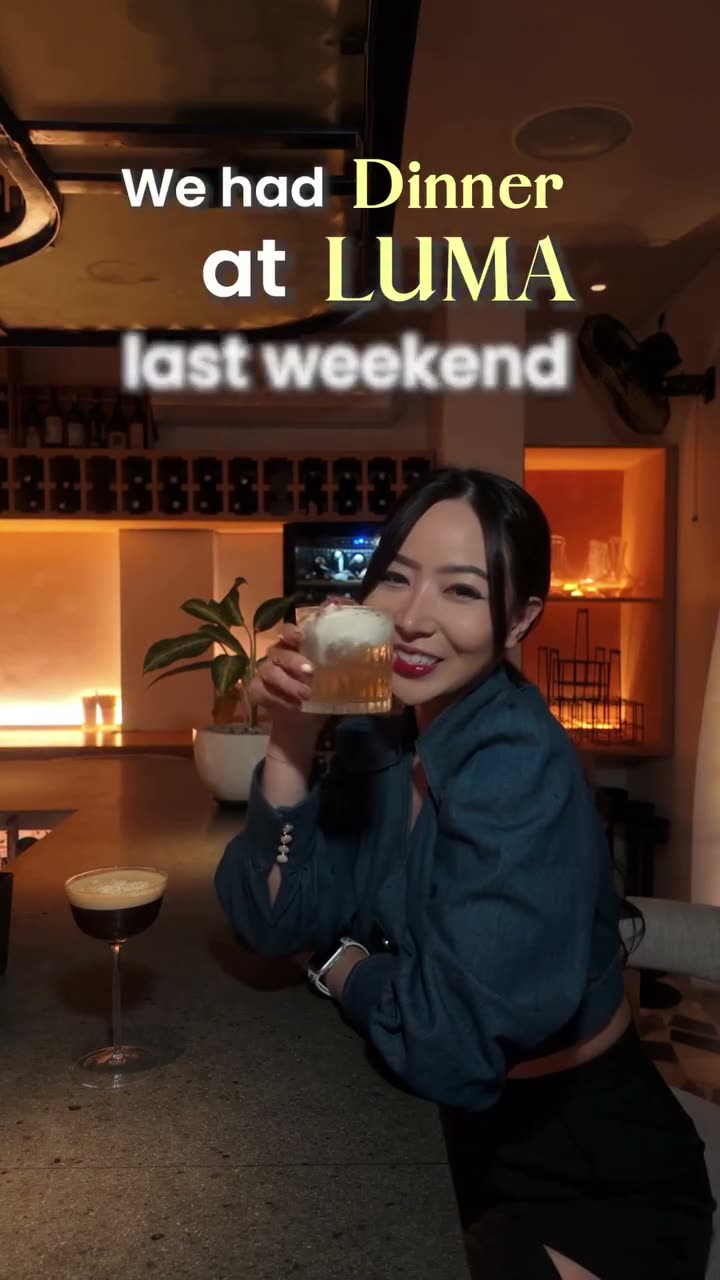 A guest at Luma's bar holding a cocktail, the lit back bar behind her