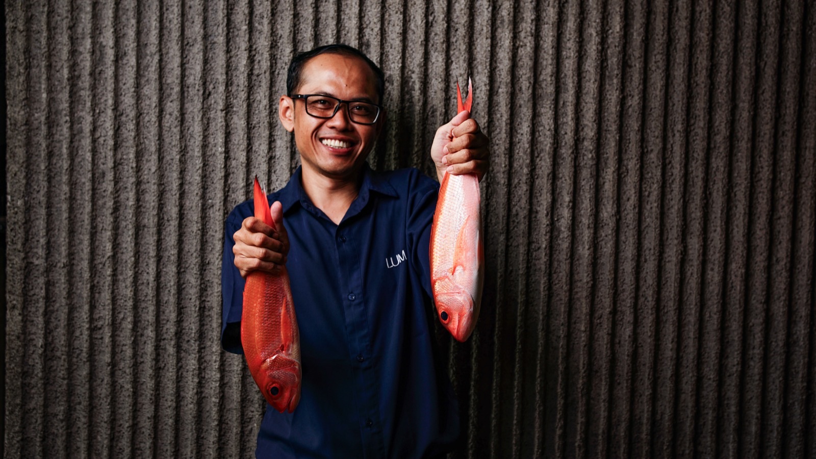 A member of Luma's team holding up two red snapper against the ribbed concrete wall